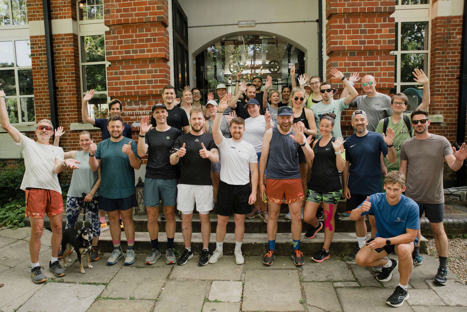 A group of people, dressed in casual sportswear, poses together cheerfully outdoors in front of a brick building. Some are raising their arms and smiling, while a dog stands on the left. The atmosphere is lively and friendly.
