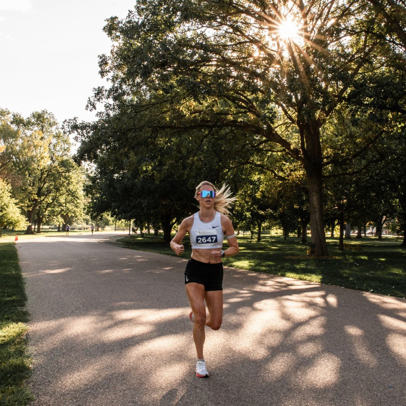 A runner wearing sunglasses and bib number 2647 jogs on a sunlit park path, surrounded by tall trees and dappled sunlight filtering through the branches.
