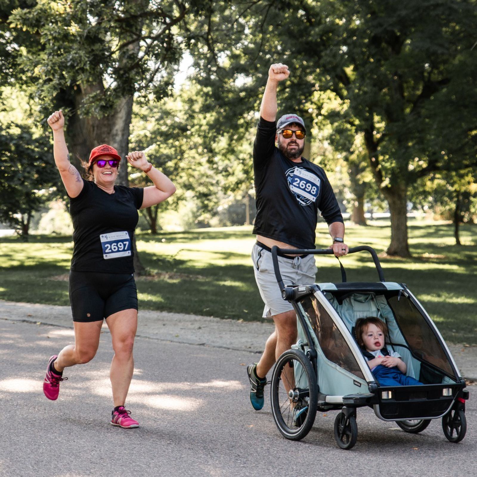 A woman and a man, both wearing race bibs, run on a park path with their arms raised. The man pushes a stroller with a young child inside. Trees and grass are in the background.
