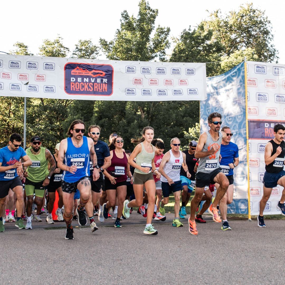 A group of runners starts a race under a banner reading βDenver Rocks Run.β The diverse group wears race bibs and athletic gear, running on a paved road with trees in the background.