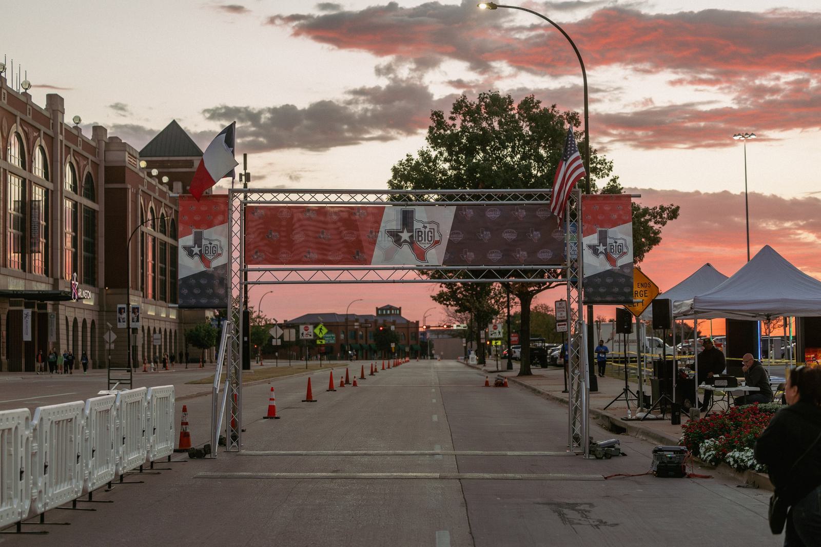 A race finish line is set up on a city street at sunrise, with American and Iowan flags on the metal archway. White barricades, orange cones, and tents line the road, and a few people are present on the right side.