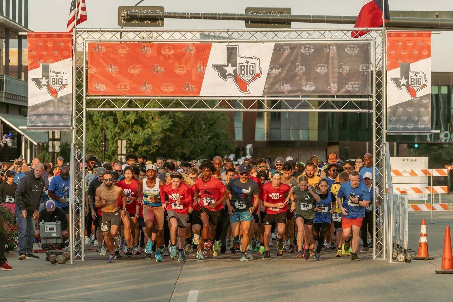 A large group of runners begins a race under a banner reading "The Big Texas," with Texas-themed graphics and flags overhead on a sunny day. Spectators and volunteers stand on the sides.