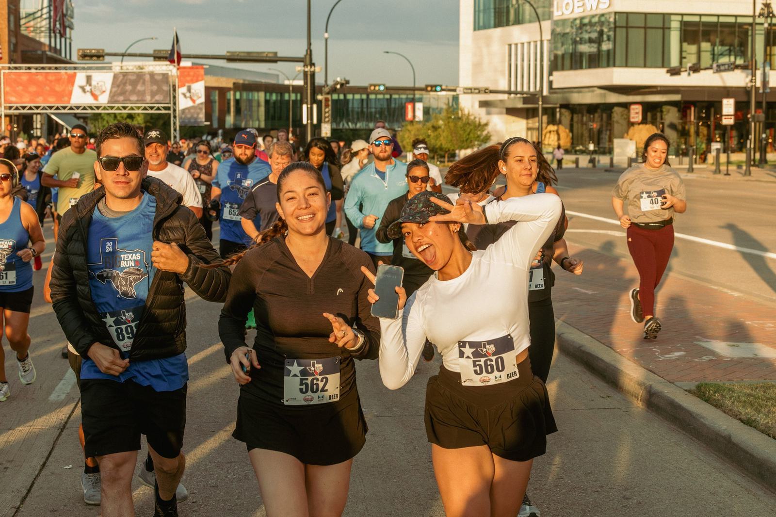Runners in athletic gear participate in a race on a city street; two women in the front smile and gesture excitedly toward the camera, while other runners follow behind in the early morning light.