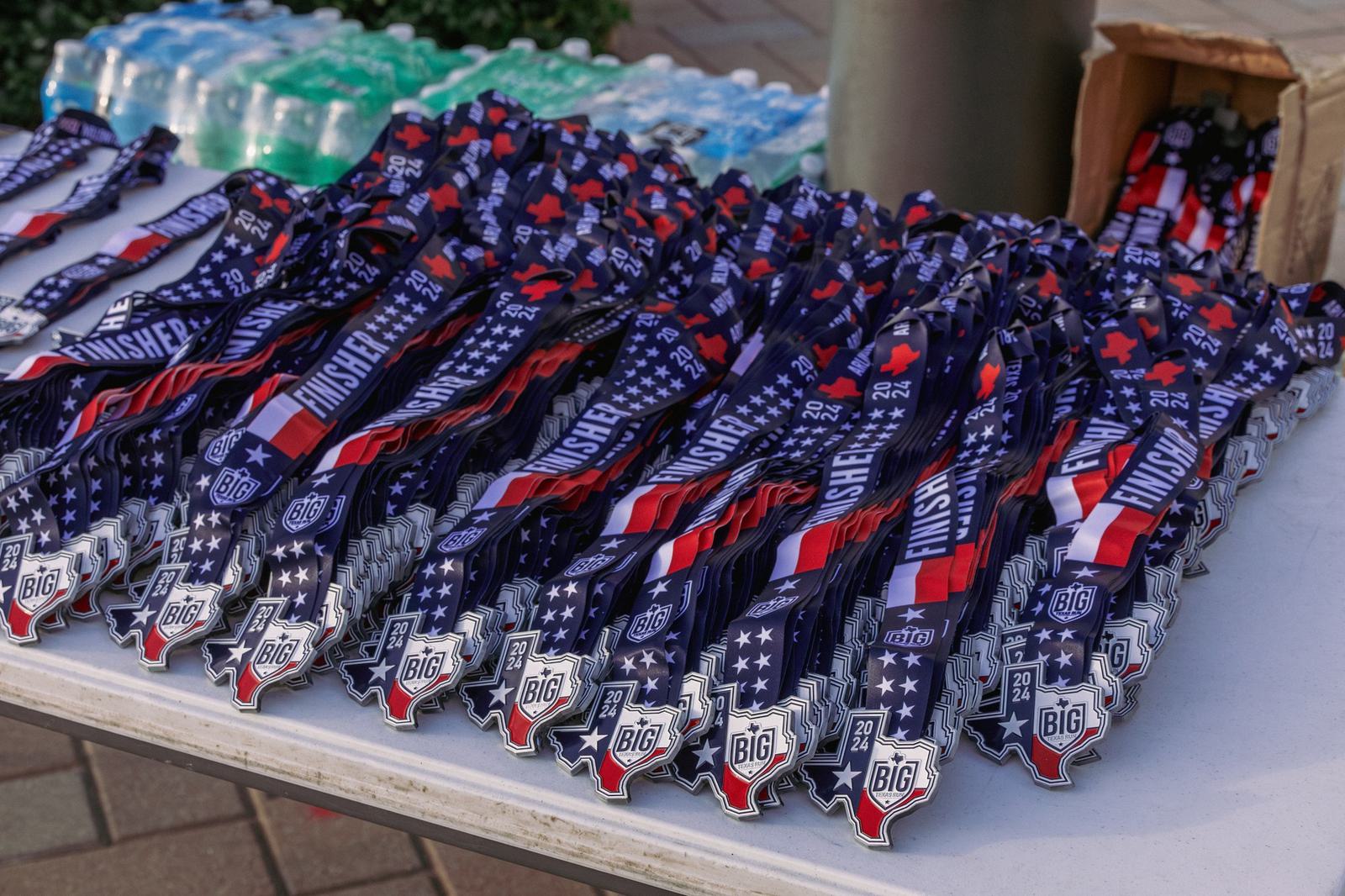 A table covered with stacks of "Finisher" medals on blue, red, and white ribbons, featuring Texas state outlines, set up outdoors next to water bottles and a cardboard box.