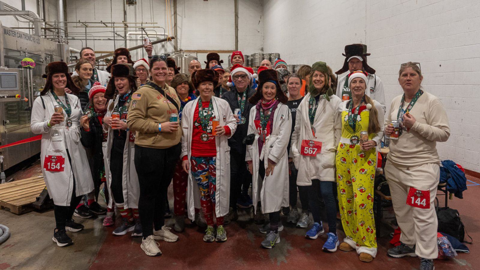 A group of people in festive clothing, hats, and costumes pose together indoors. Many wear white coats, medals, and race bibs, standing closely and smiling for the photo in an industrial setting.