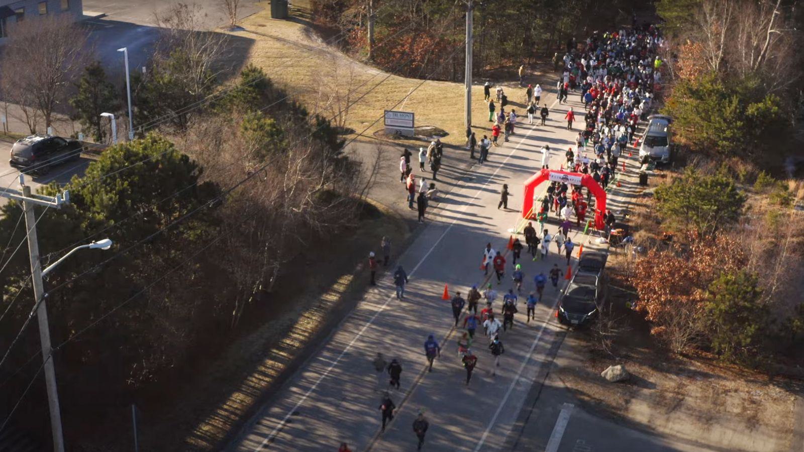 Aerial view of a road race with runners passing under a red inflatable arch, surrounded by trees and spectators along the sides on a sunny day.