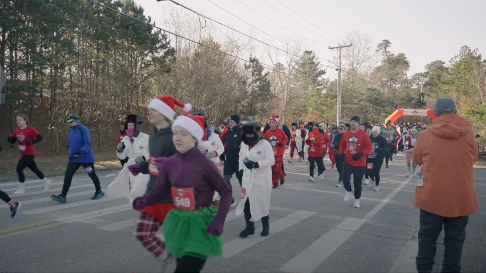 A group of people dressed in festive holiday outfits, including Santa hats, costumes, and running gear, participate in a road race on a chilly day with trees and an inflatable finish line in the background.