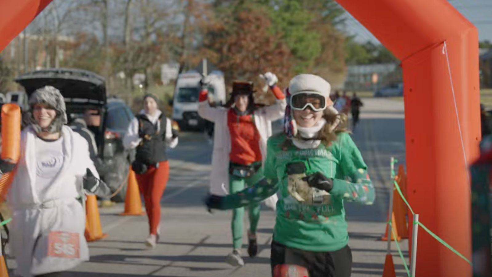 A group of runners wearing festive costumes and winter hats cross under an orange inflatable race arch, smiling and celebrating at an outdoor event on a sunny day.