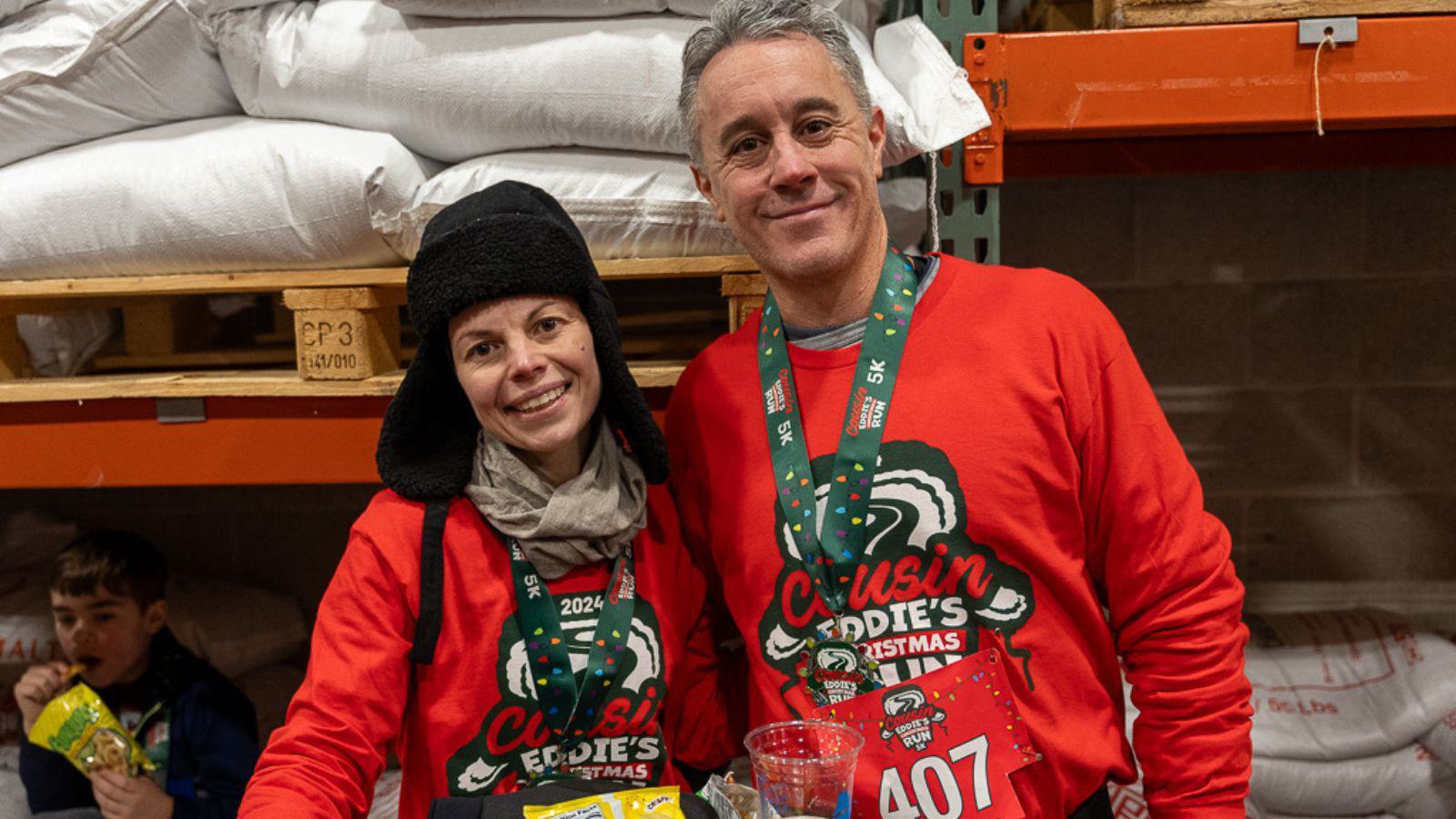 Two people wearing red "Cousin Eddie's Christmas" shirts and medals stand smiling together indoors by shelves with bags. One wears a black winter hat; both have race bibs. A child is in the background eating a snack.