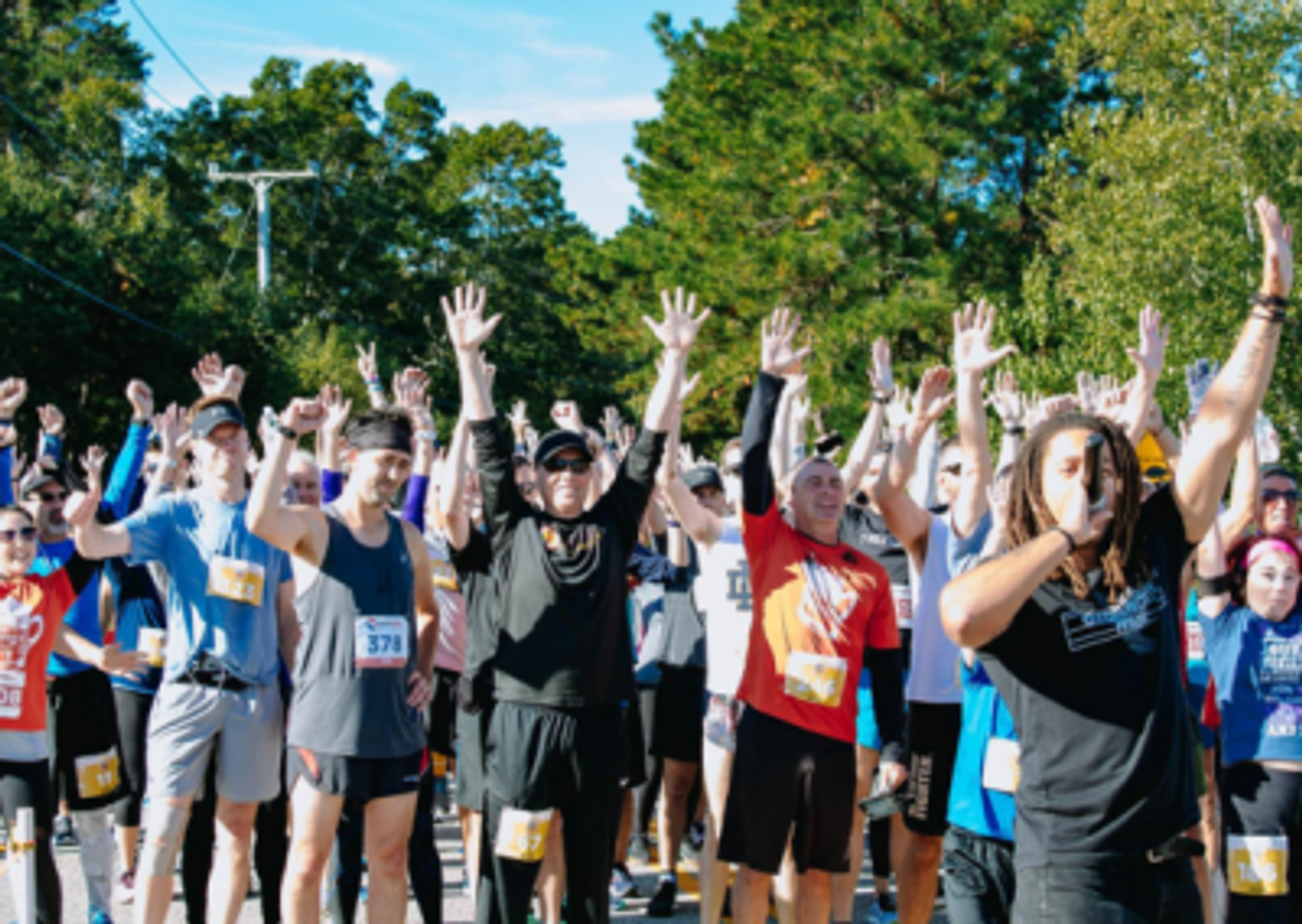 A large group of runners stand outdoors with their hands raised, some smiling, at the starting line of a race on a sunny day, surrounded by trees. An announcer with a microphone is visible in the foreground.