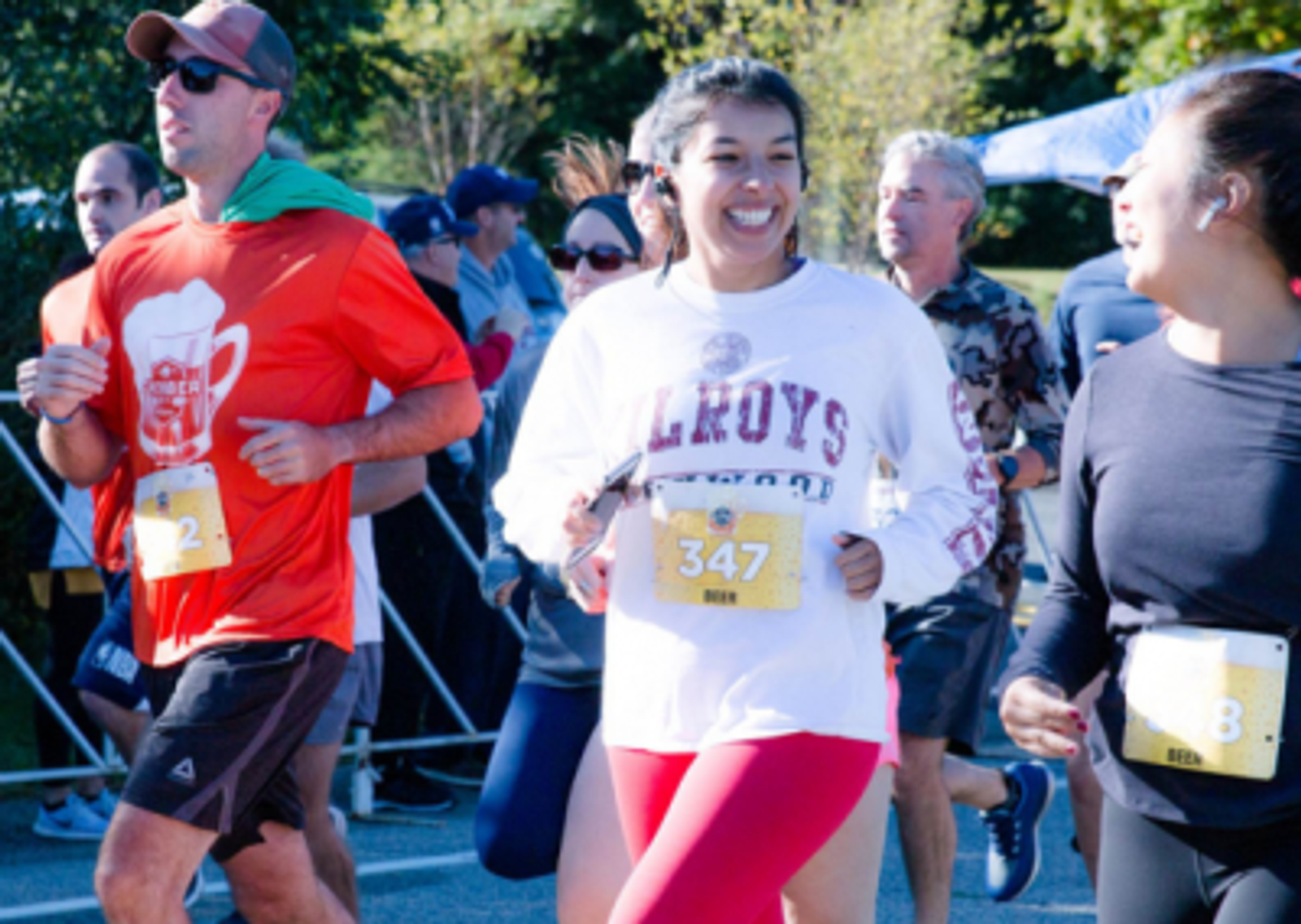 A group of runners, wearing race bibs and casual athletic outfits, smile as they participate in an outdoor race on a sunny day. Trees and other runners are visible in the background.