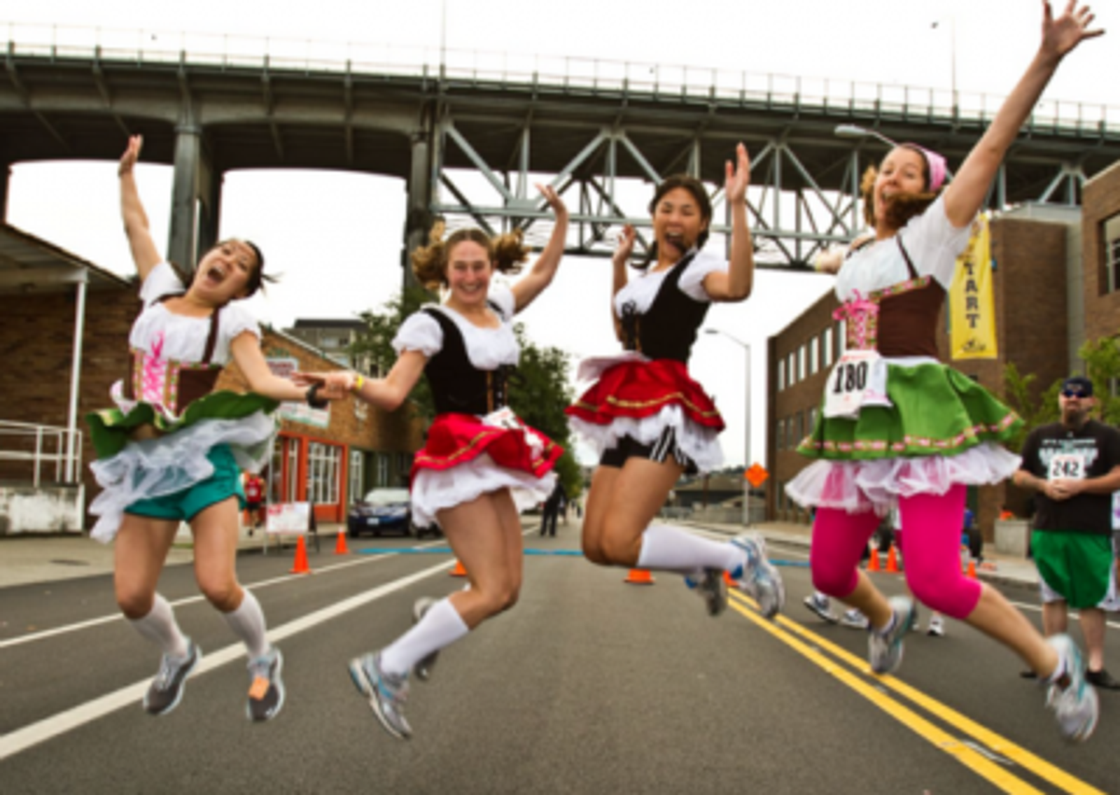 Four women in colorful costumes jump in the air, smiling, during a street race. They are under a bridge, and other runners and buildings are visible in the background.