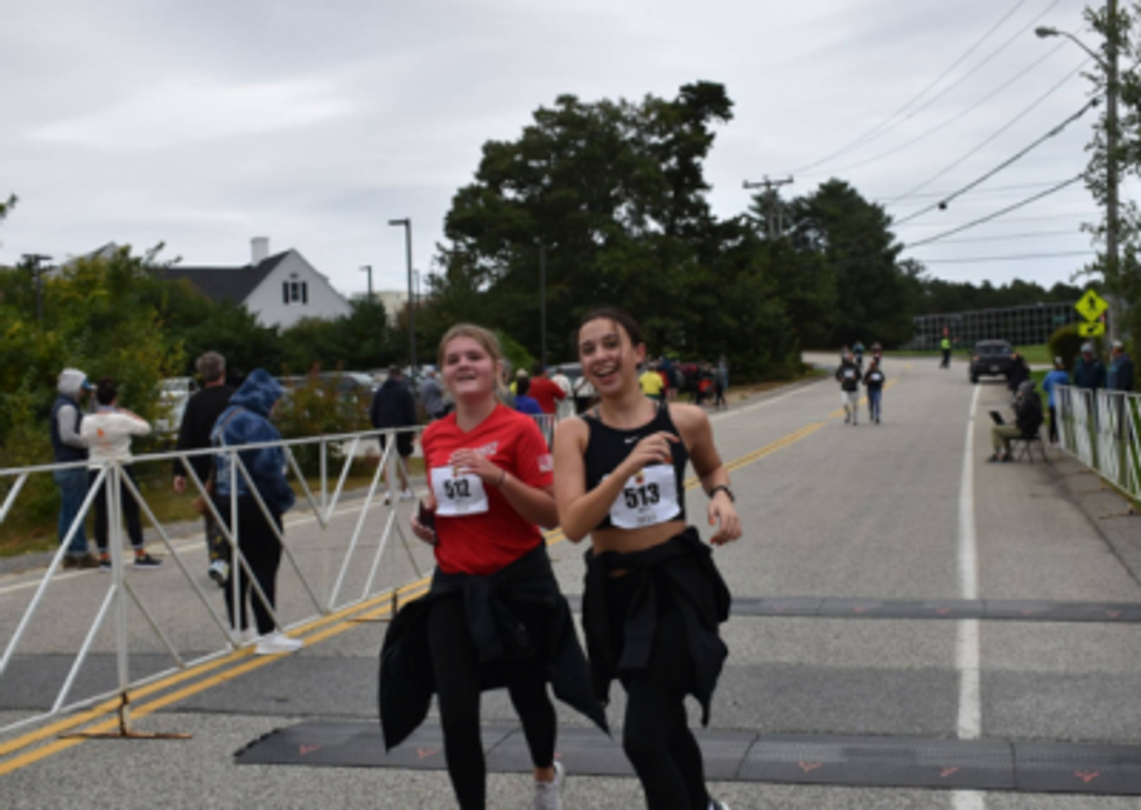 Two women wearing race bibs and athletic clothes smile as they cross a race finish line together on a road, with spectators and trees in the background.