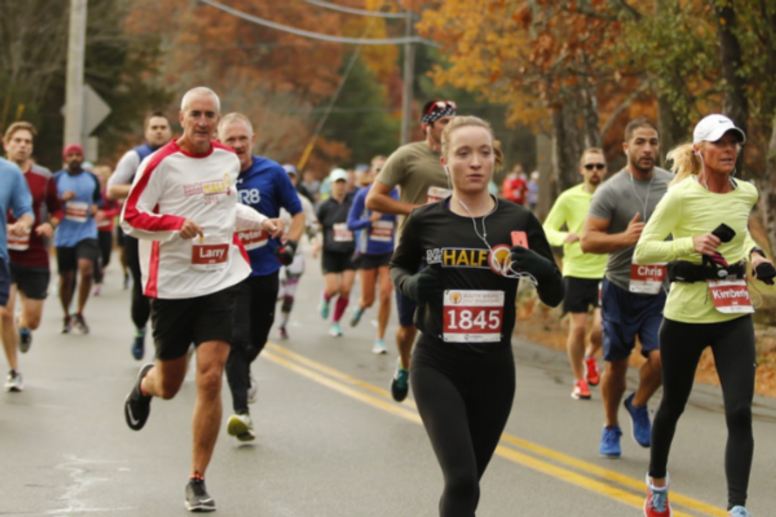 A group of runners participates in a road race on an overcast day. The runners are wearing bib numbers and athletic gear, with autumn trees and a street in the background.