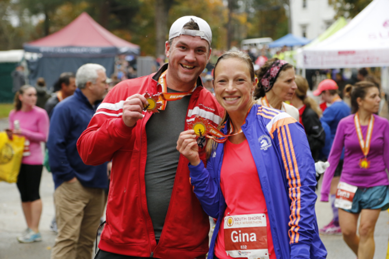 Two runners smiling and holding up medals after a race. The woman wears a purple jacket and has a race bib labeled "Gina." The man wears a red jacket. Other participants and tents are visible in the background.