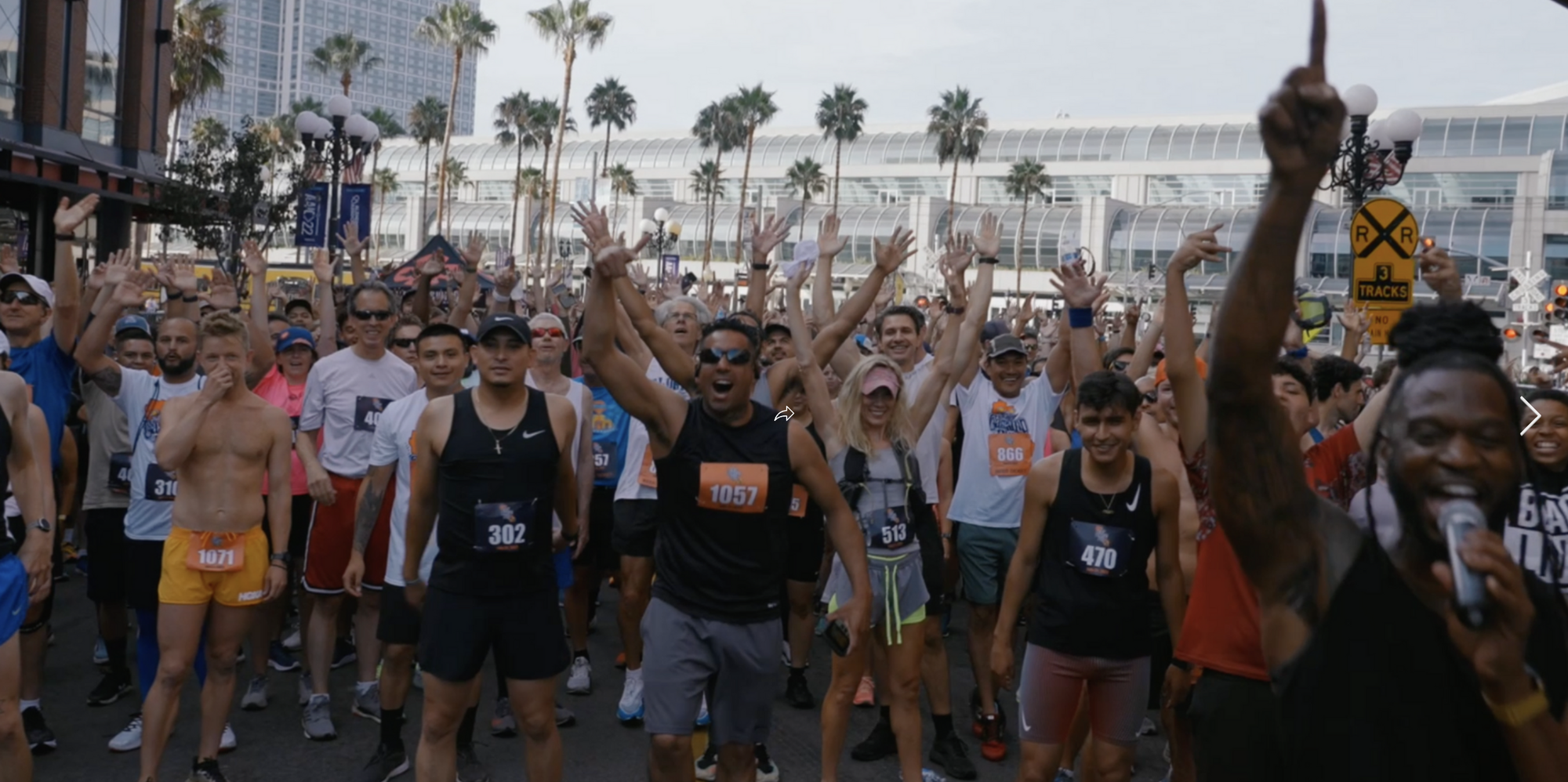 A large, cheerful crowd of runners gathers at the starting line of a race. Many have their hands raised and are smiling, showing excitement. An energetic person with a microphone is at the right side of the image, leading the group. Palm trees and modern buildings are in the background.