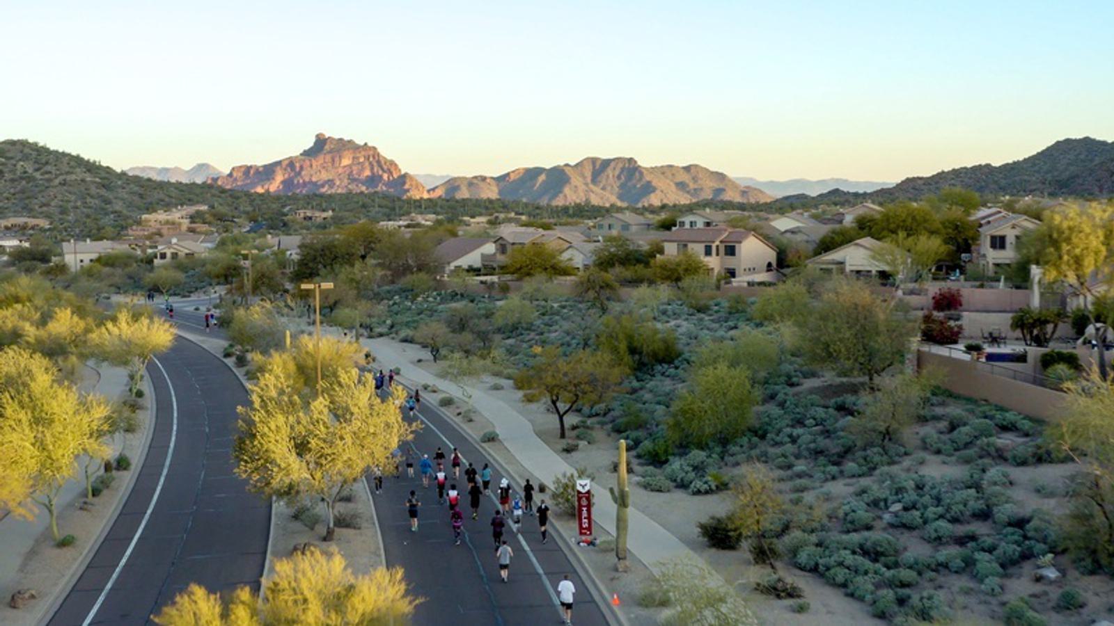 A group of runners jogs down a paved road surrounded by desert landscape and residential homes. In the background, rugged mountains rise under a clear sky at sunset. Vegetation typical of a desert environment can be seen on either side of the road.