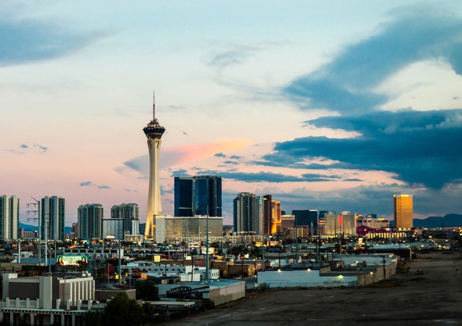 A panoramic view of a city skyline at sunset, featuring a prominent tower with a spherical top. Surrounding the tower are various high-rise buildings with reflective glass windows. The sky is a mixture of soft blues, pinks, and oranges, with scattered clouds.