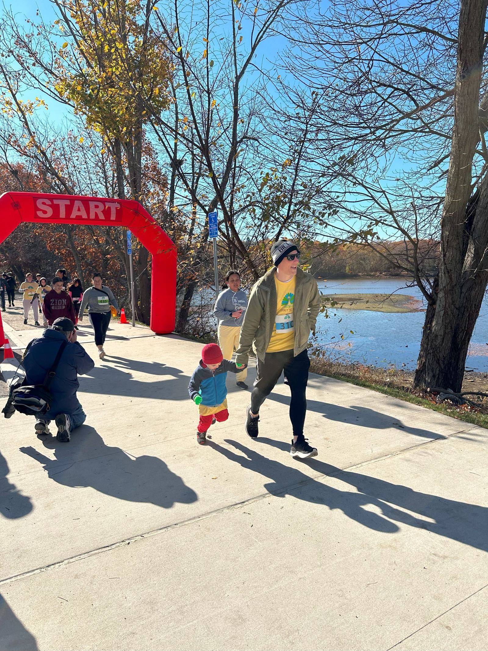 People walk past a red "START" arch at an outdoor event near a river on a sunny day. A child in a red hat and an adult in a green jacket are in the foreground, with others and trees in the background.