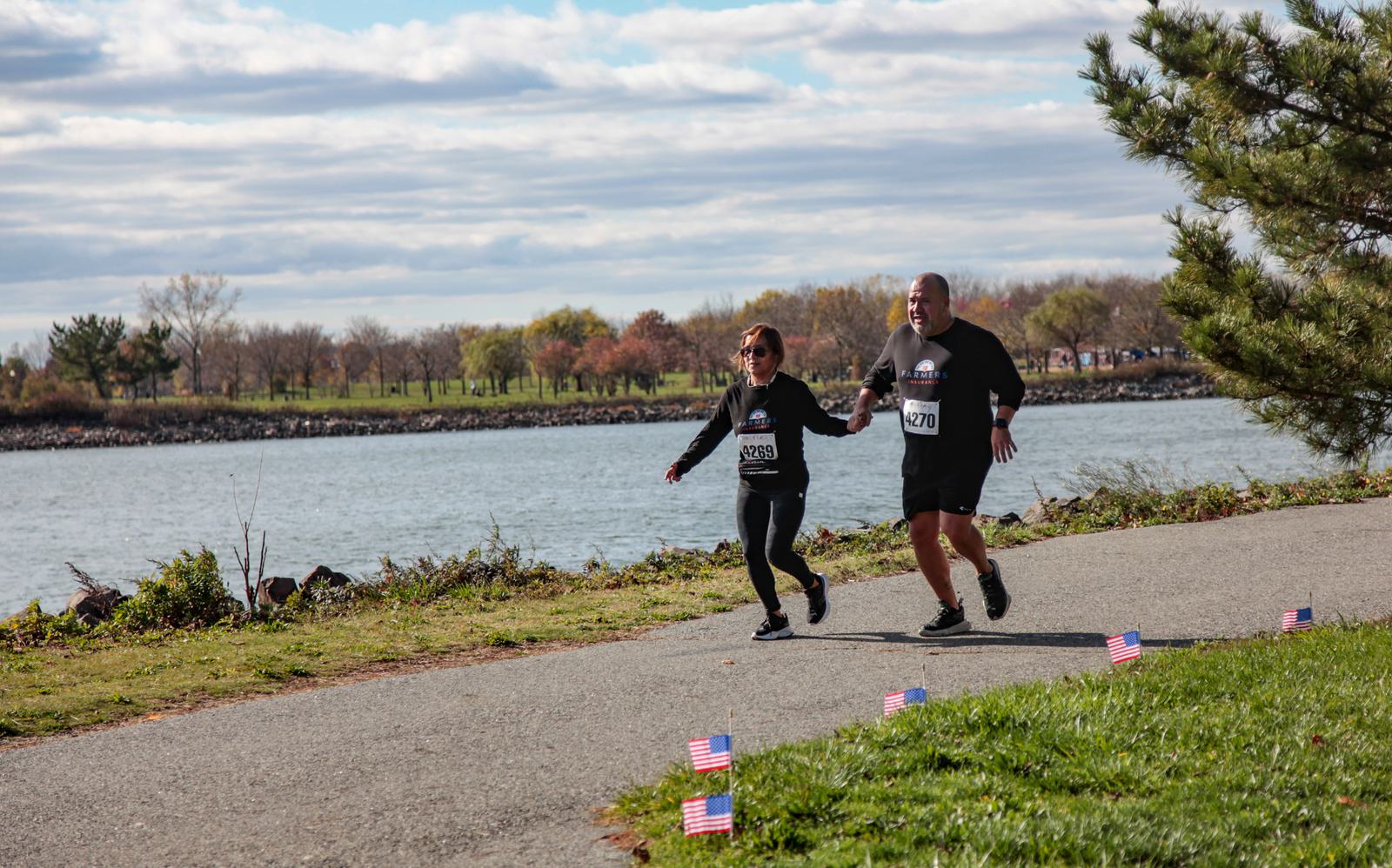 Two people in athletic wear are jogging on a paved path beside a river, with small American flags lining the trail. The sky is partially cloudy, and trees are visible in the background.