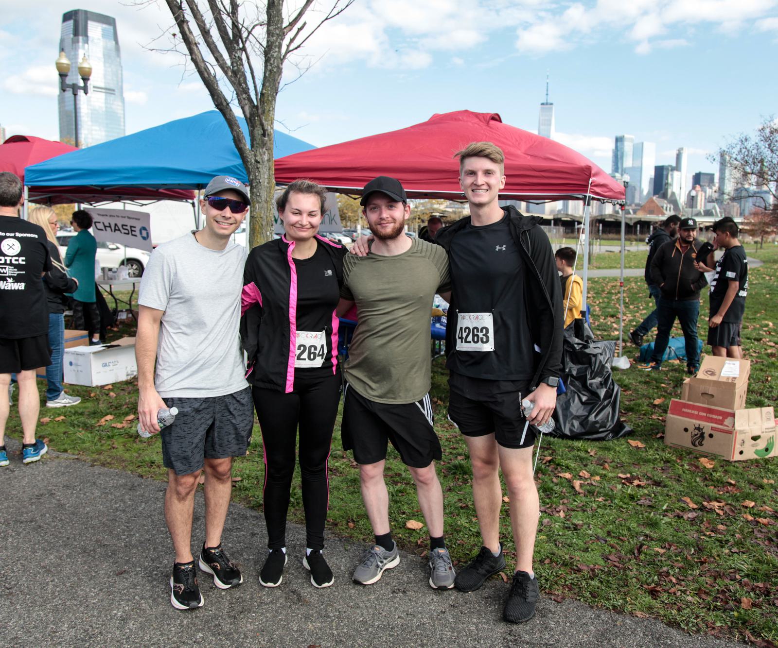 Four people stand together after a race, smiling for a photo. They wear athletic gear with race numbers. Tents and a city skyline are visible in the background.