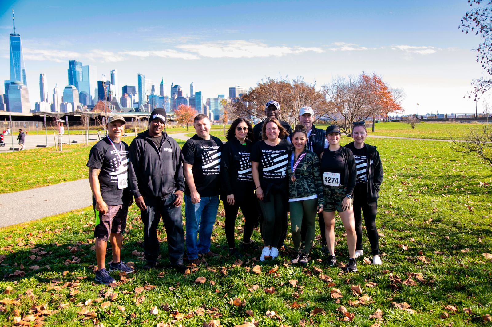 A group of people in matching shirts pose for a photo in a park with a city skyline in the background. The grass is dotted with fallen leaves, and a few trees are visible. One person wears a bib number. The sky is clear and blue.