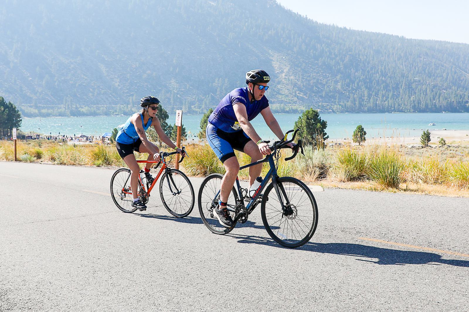 Two cyclists ride on a scenic road near a lake surrounded by mountains. The leading cyclist wears a blue outfit, while the other wears a black and blue ensemble. They are biking under clear skies with a view of distant trees and hills.