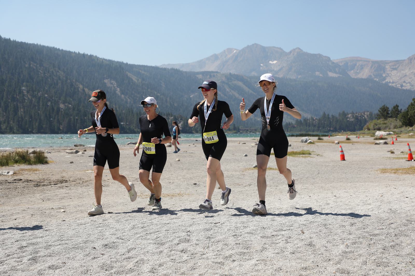 Four women in athletic wear run on a sandy trail near a lake, with medals around their necks. They are smiling and giving thumbs up against a backdrop of mountains and blue sky. Orange traffic cones line the trail.