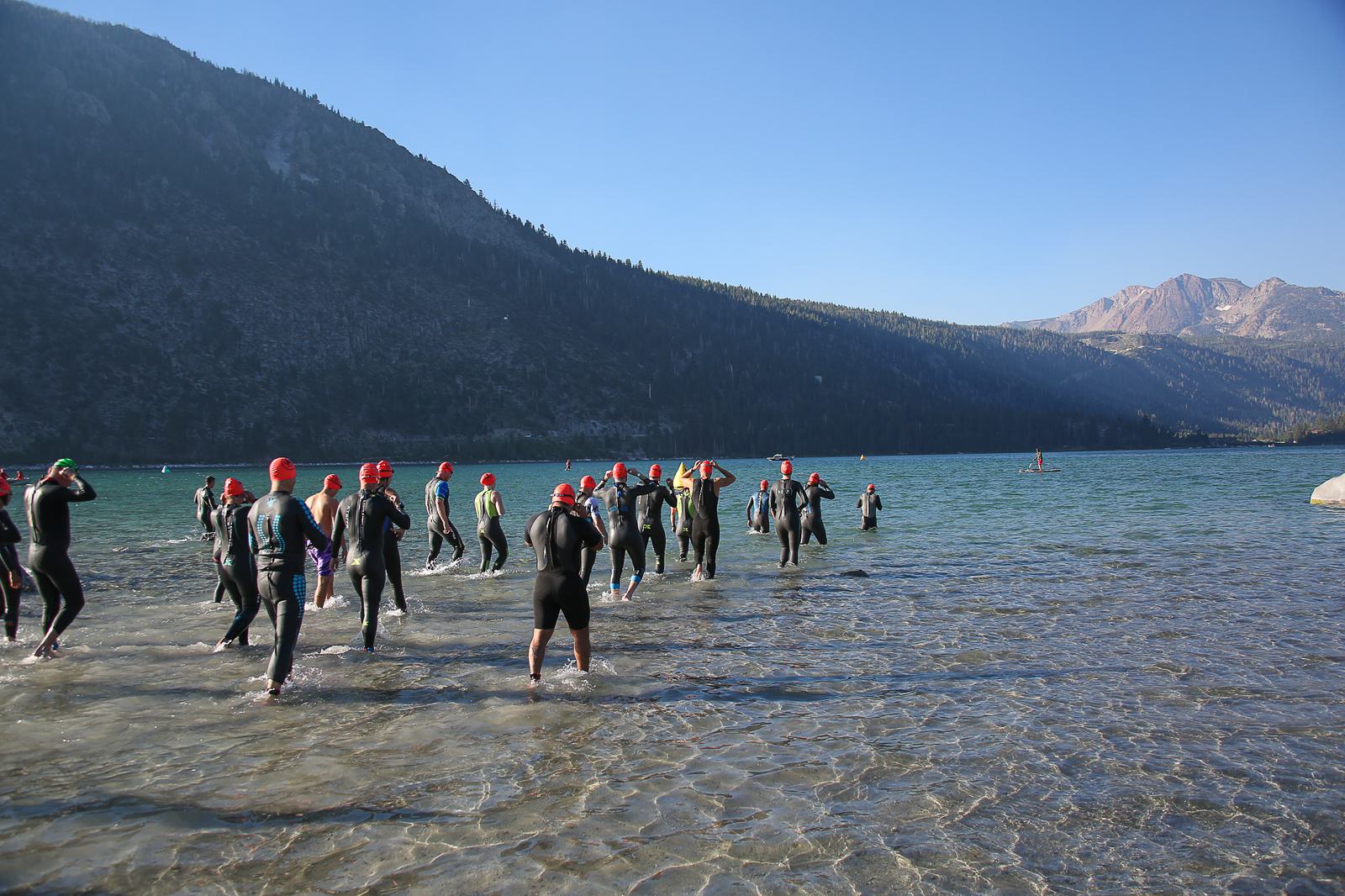 A group of people wearing wetsuits and red swim caps enter a clear mountain lake with distant forested hills and blue sky. They appear to be preparing for a swim or race.