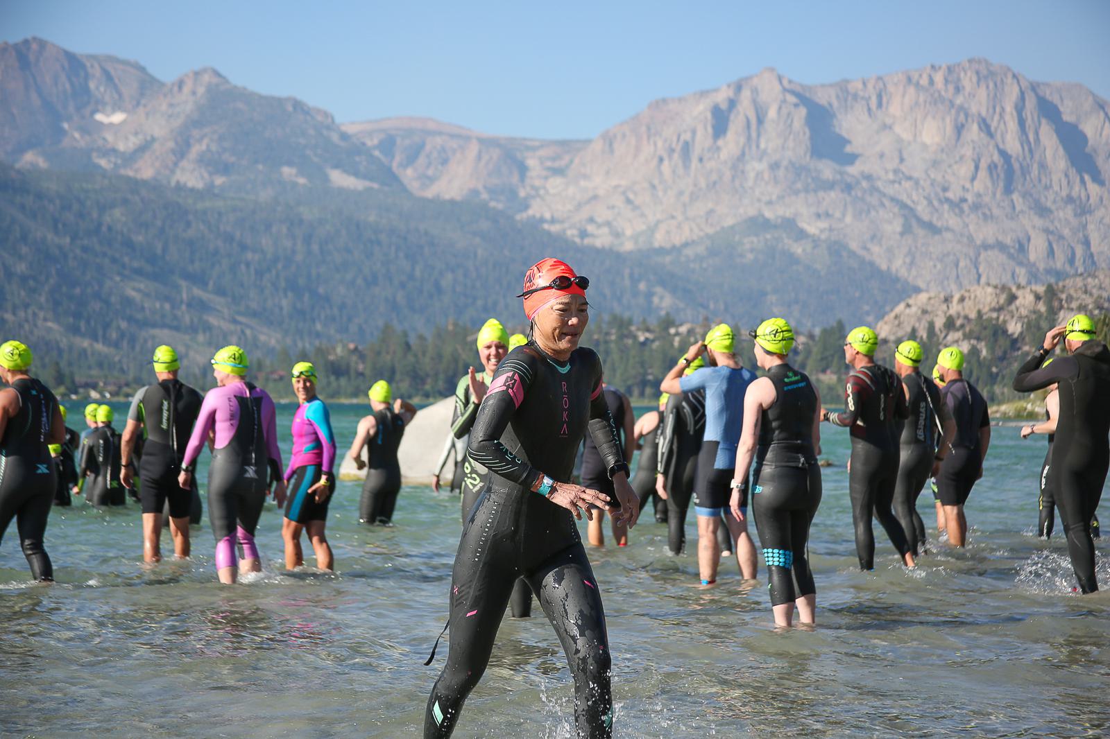 Participants wearing wetsuits and swim caps begin a swim race in a lake surrounded by mountains. One person in a red cap is in the foreground, exiting the water, while others follow behind. The sky is clear and blue.