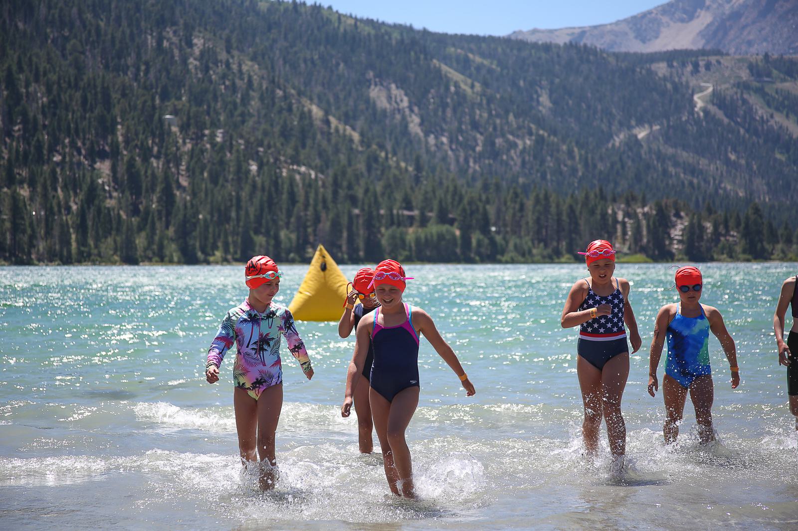 A group of children wearing swim caps wade through a clear blue lake. They are participating in a swimming event, with a buoy visible in the water. In the background, there are forested mountains under a clear sky.