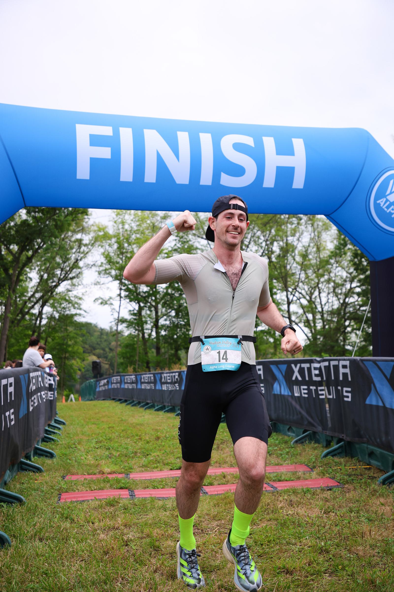 A runner in athletic gear crosses a blue finish line arch, displaying a triumphant fist pump. The man wears a cap backward, a light gray top, black shorts, and bright yellow-green socks. Greenery and branding banners line the grassy path.