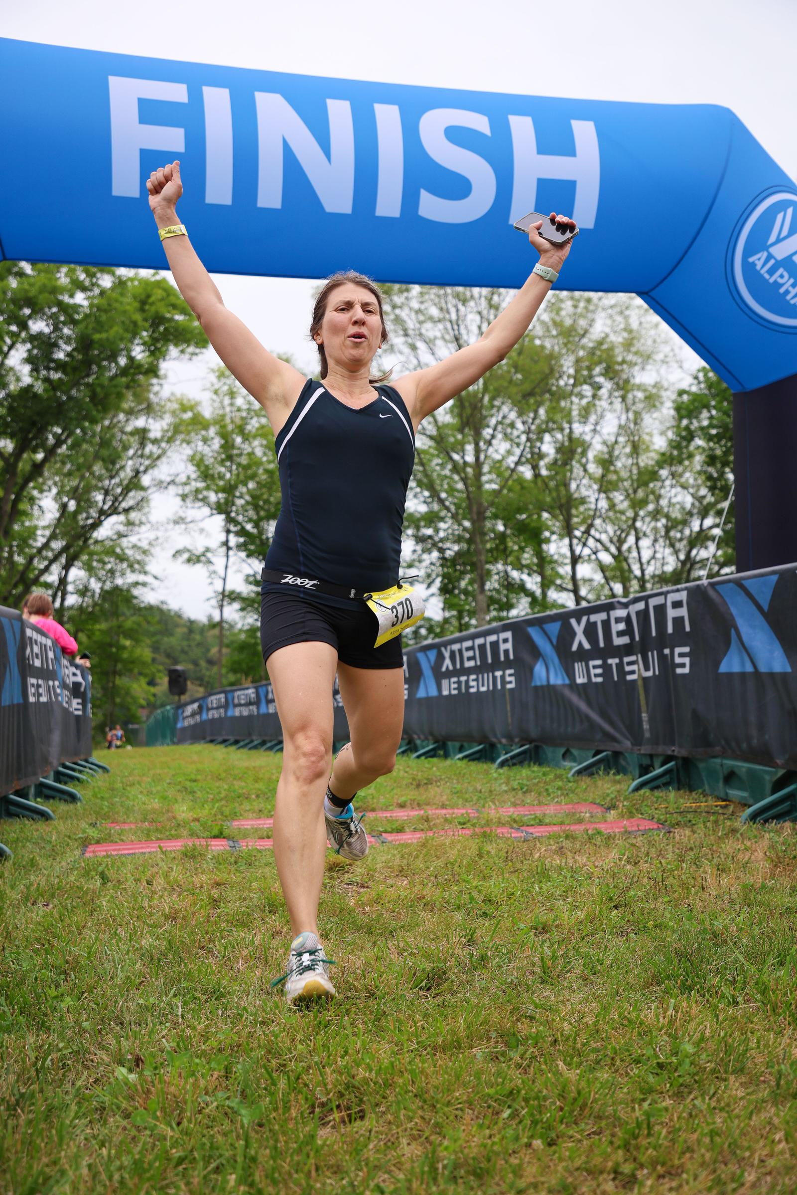 A woman in athletic wear crosses the finish line of a race, holding a phone and raising one arm in triumph. The finish line banner and race barriers are visible, with a grassy setting and trees in the background.