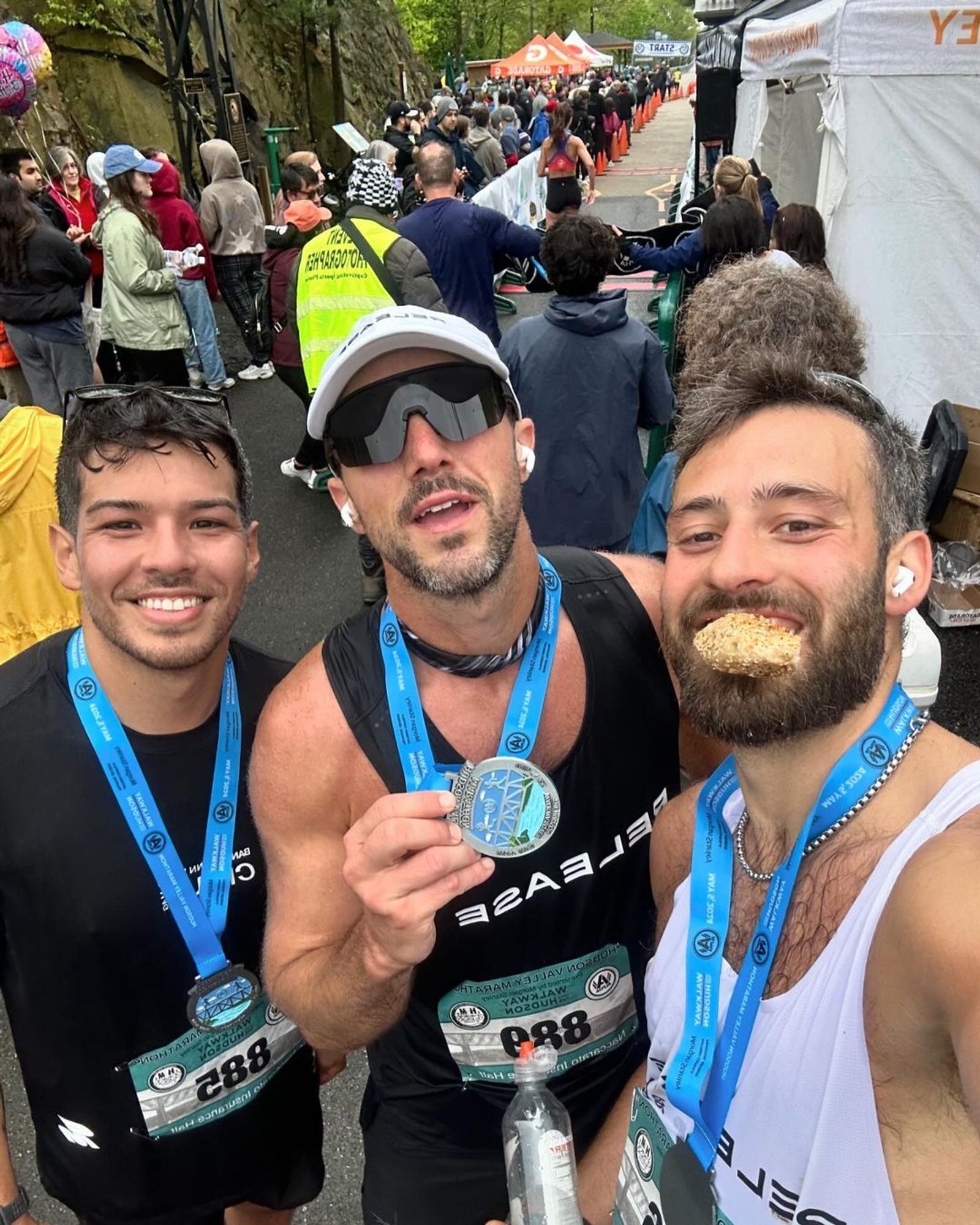 Three men with medals around their necks pose for a photo after a race. One man is holding up his medal, while another is taking a bite of a snack. They are surrounded by other participants and tents in the background.