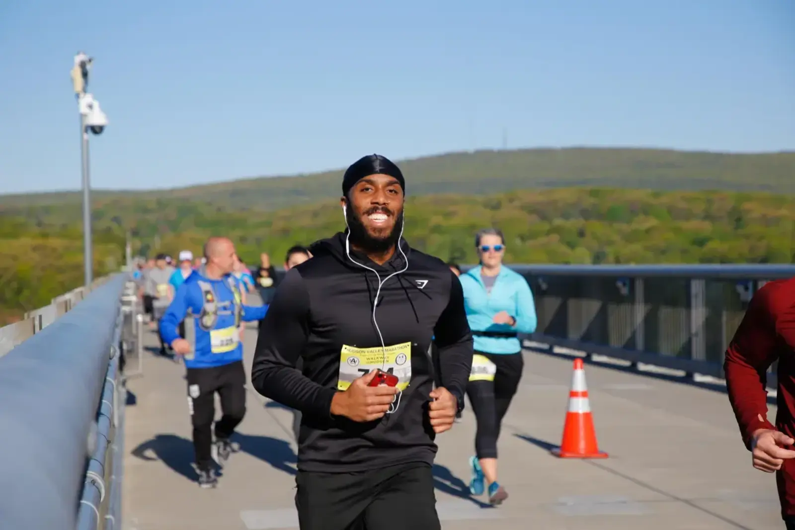 A group of people running in a race on a bridge under a clear blue sky. A person in the foreground is smiling, wearing a black headband and hoodie. Participants wear bib numbers. Green hills are visible in the background.
