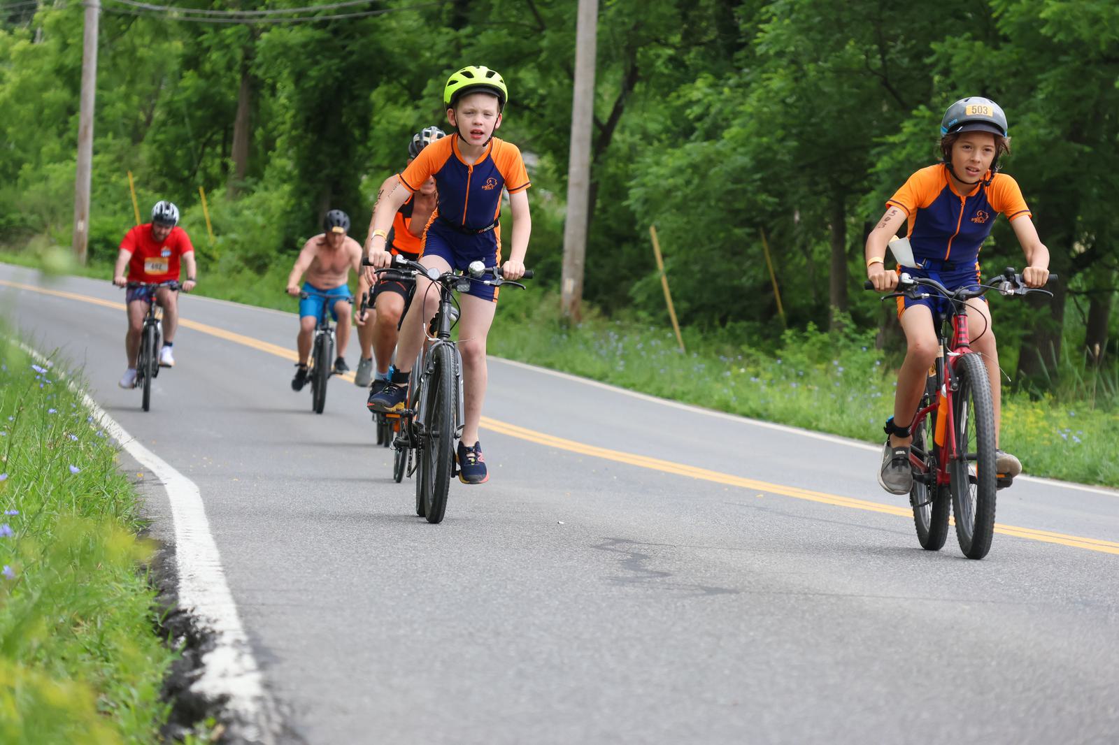 A group of cyclists rides on a rural road lined with trees. Two young cyclists in orange jerseys lead, while others follow behind. The sky is clear, and wildflowers grow along the roadside.