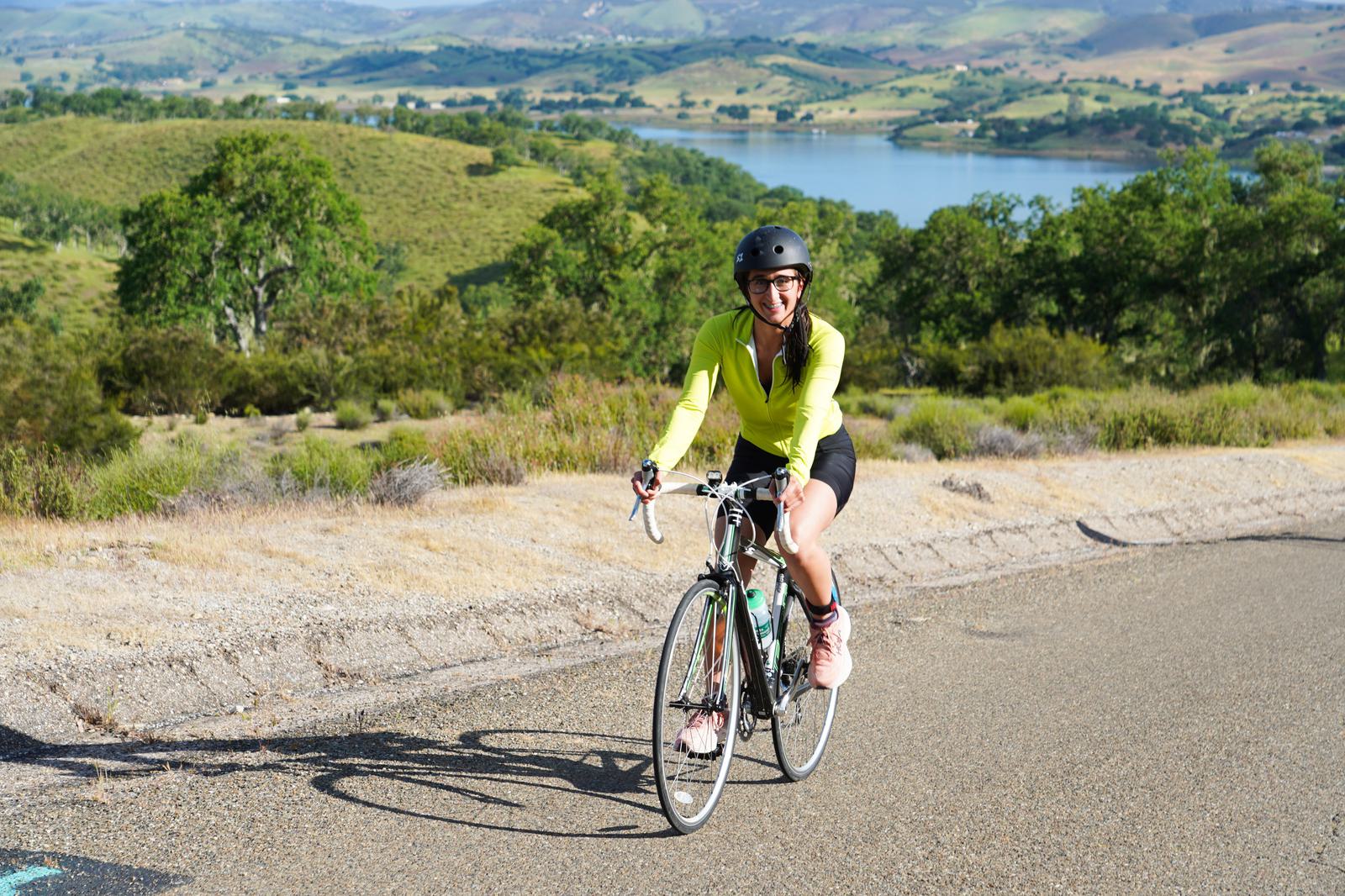 A person wearing a helmet and bright green jacket rides a bicycle on a paved road. The scenic background features lush green hills and a large lake under a clear blue sky.
