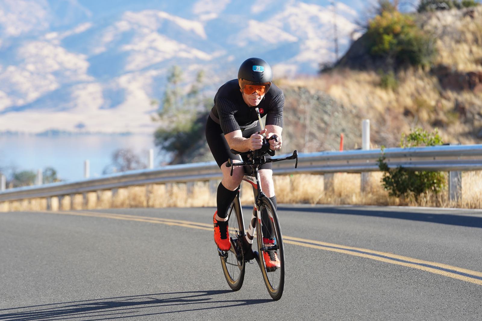 A cyclist in black gear and a helmet rides a road bike on a sunny mountain road. The landscape features dry grass, hills, and a clear blue sky. The cyclist is focused and in a crouched racing position.