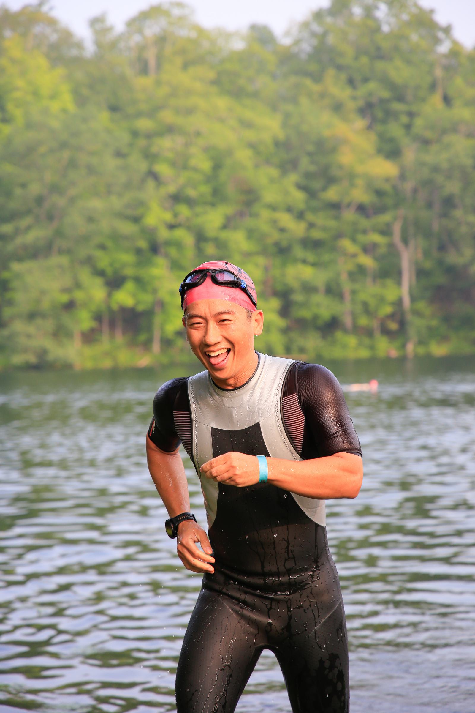 A person wearing a wetsuit and pink swim cap is running out of a lake, smiling and appearing joyful. The background shows a forested area with lush green trees.