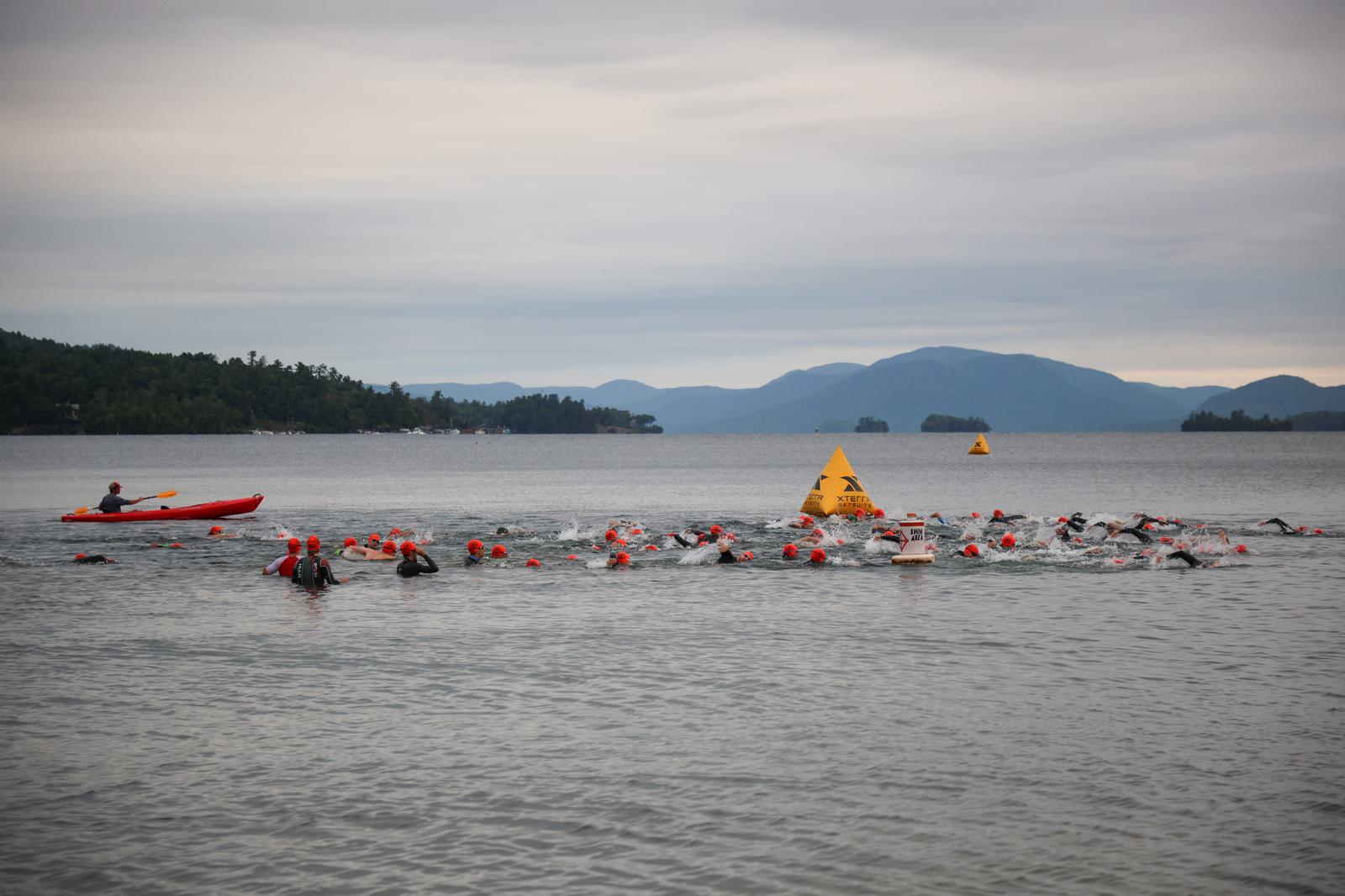 A group of swimmers in wetsuits and orange caps participate in a race in a lake. A safety kayaker follows beside them. There are orange buoys in the water, and hilly islands are visible in the background under a cloudy sky.