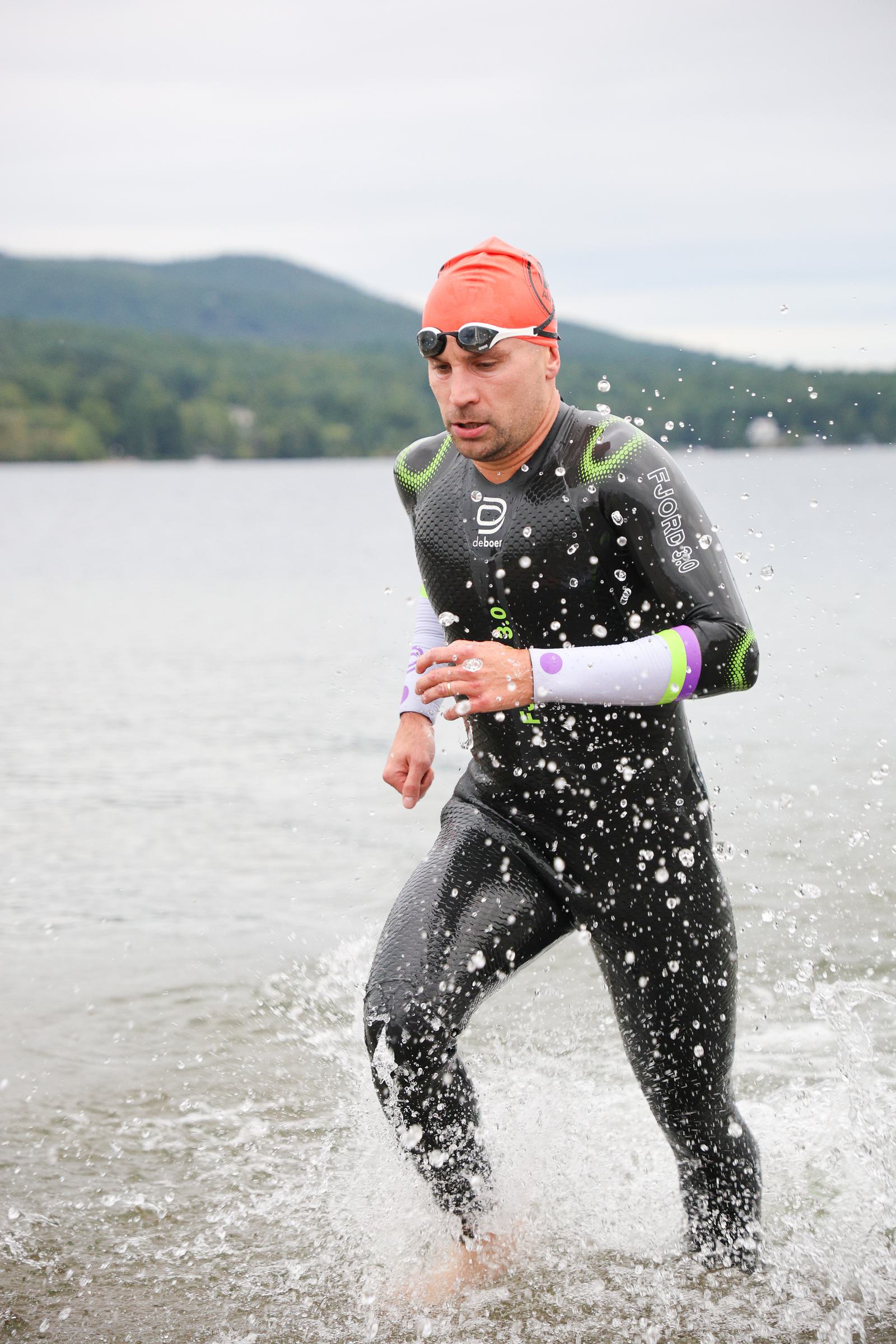 A person wearing a black wetsuit, orange swim cap, and goggles runs through shallow water with splashes around them. They are emerging from a lake with green hills and trees in the background. The sky is overcast.