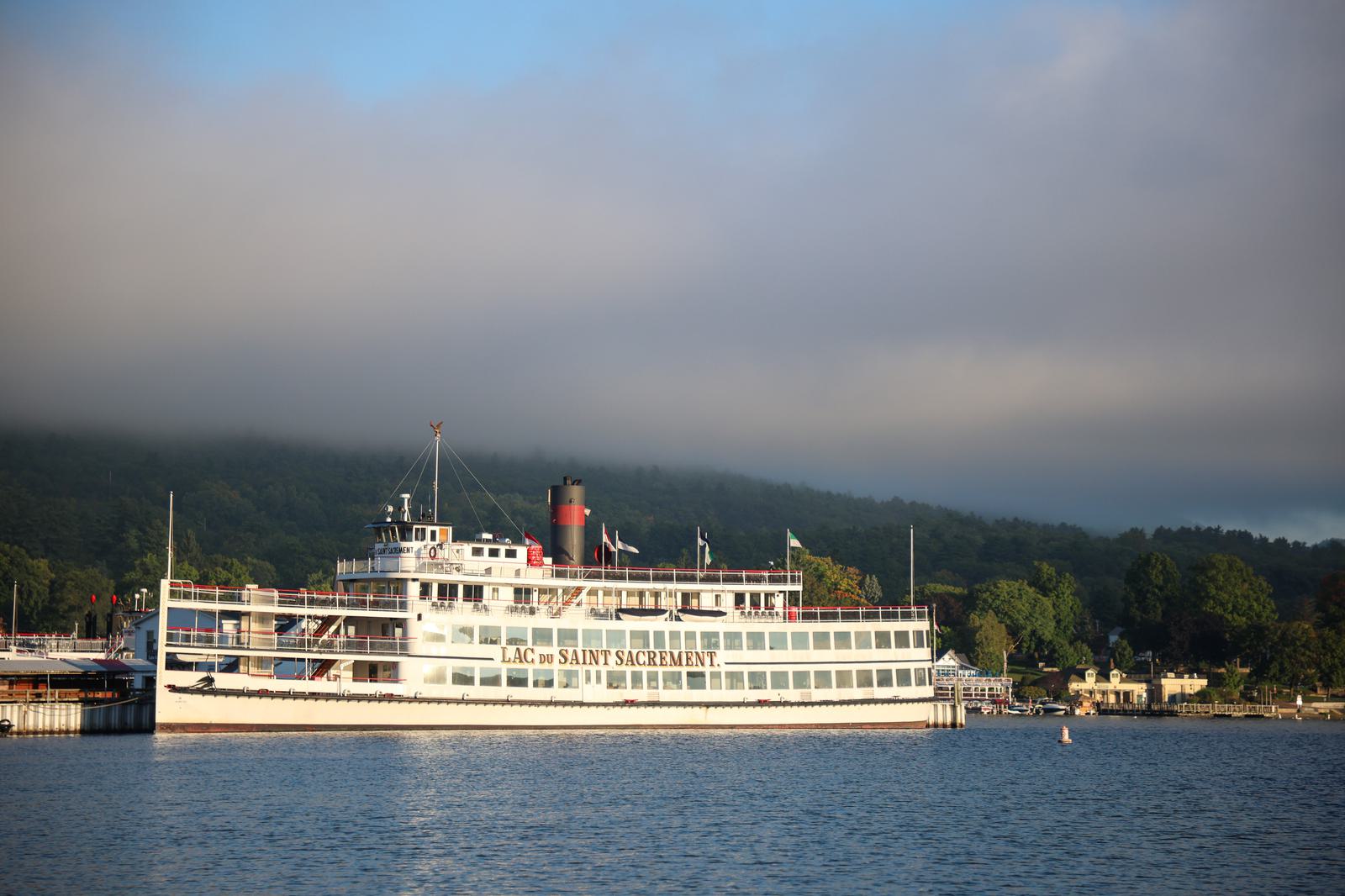 A white and red steamboat named "Lac du Saint Sacrement" is docked on a calm lake, with hills and trees in the background under a cloudy sky.