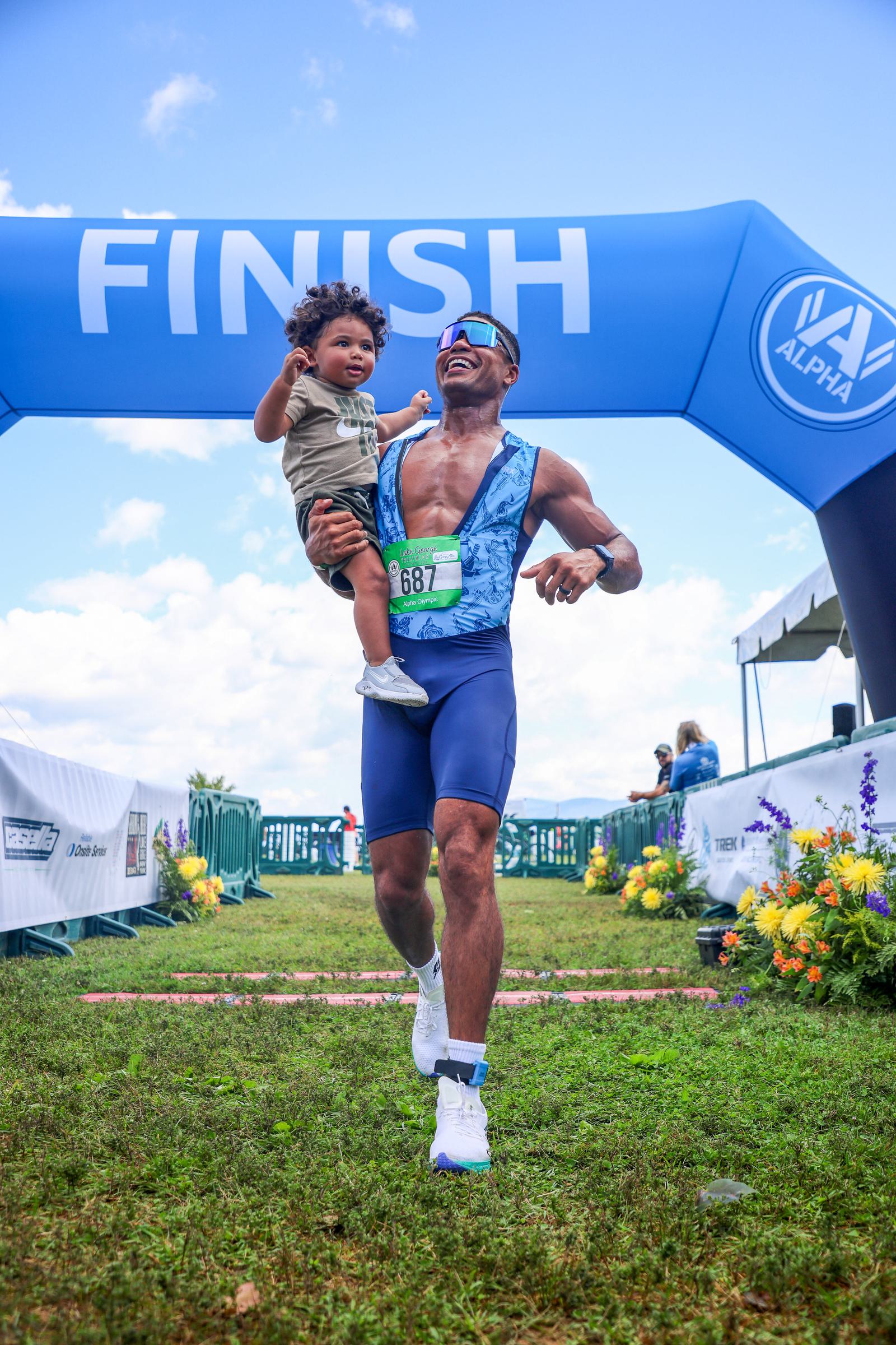 A man in athletic gear crosses a race finish line, joyfully holding a child in one arm. Both are smiling, and the child has curly hair. The finish line banner is blue, and there are flowers and tents in the background under a partly cloudy sky.