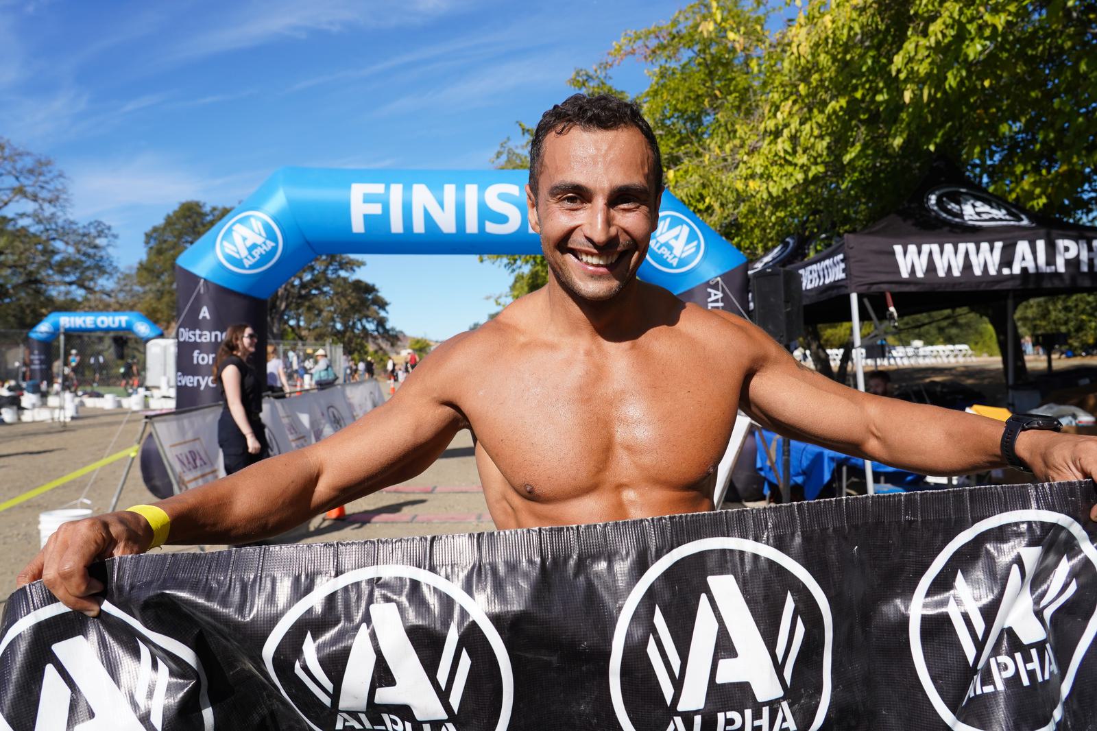 A shirtless man smiles while holding a banner at the finish line of a race. The blue and white inflatable arch behind him reads "FINISH." The race branding is visible on surrounding tents and banners.