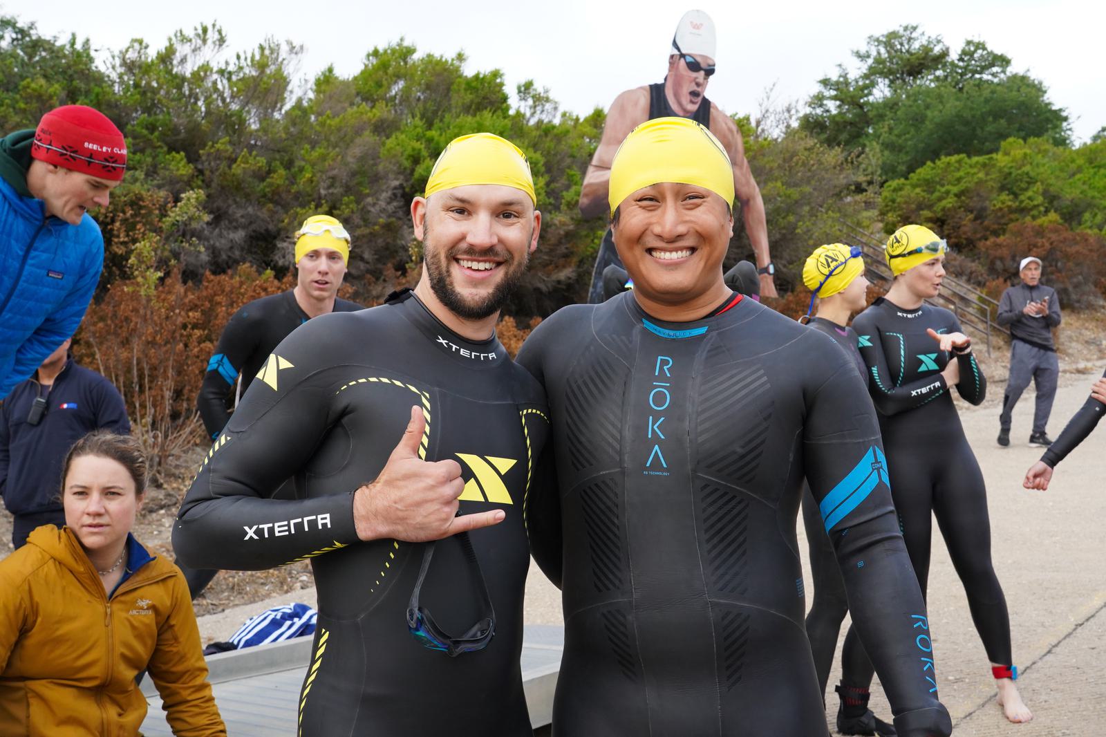 Two smiling swimmers wearing wetsuits and yellow swim caps pose together in the foreground. Several other swimmers are gathered in the background, wearing similar attire, preparing for an event in an outdoor setting with trees.