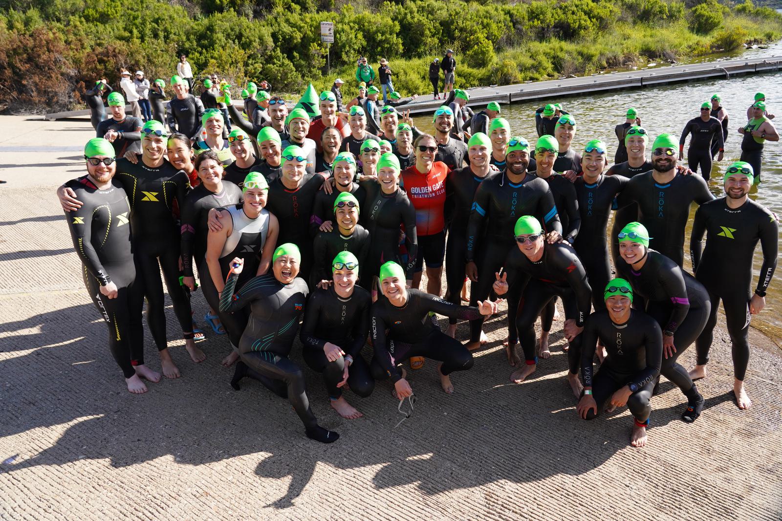 A large group of people wearing wetsuits and green swim caps gather near a waterfront for a group photo. They are smiling and posing, with greenery visible in the background. It appears to be a sunny day.