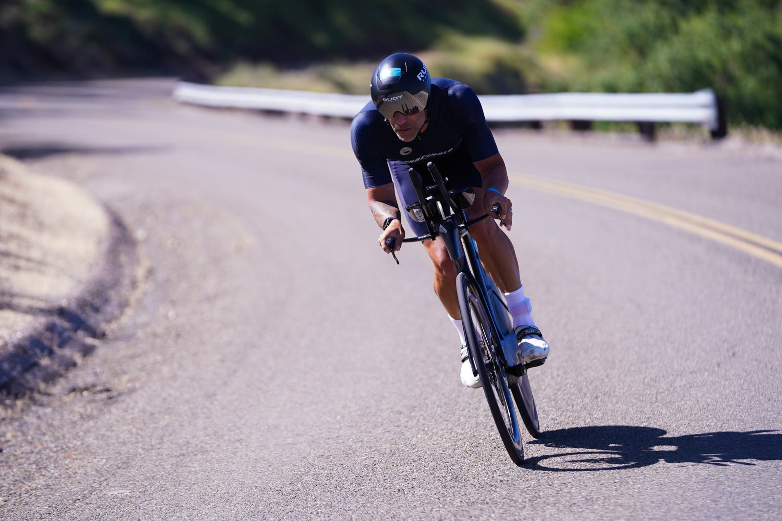 A cyclist in a black outfit and helmet leans into a curve on a sunny road, riding a high-performance bike. The background features greenery and a metal guardrail. The scene conveys speed and focus.
