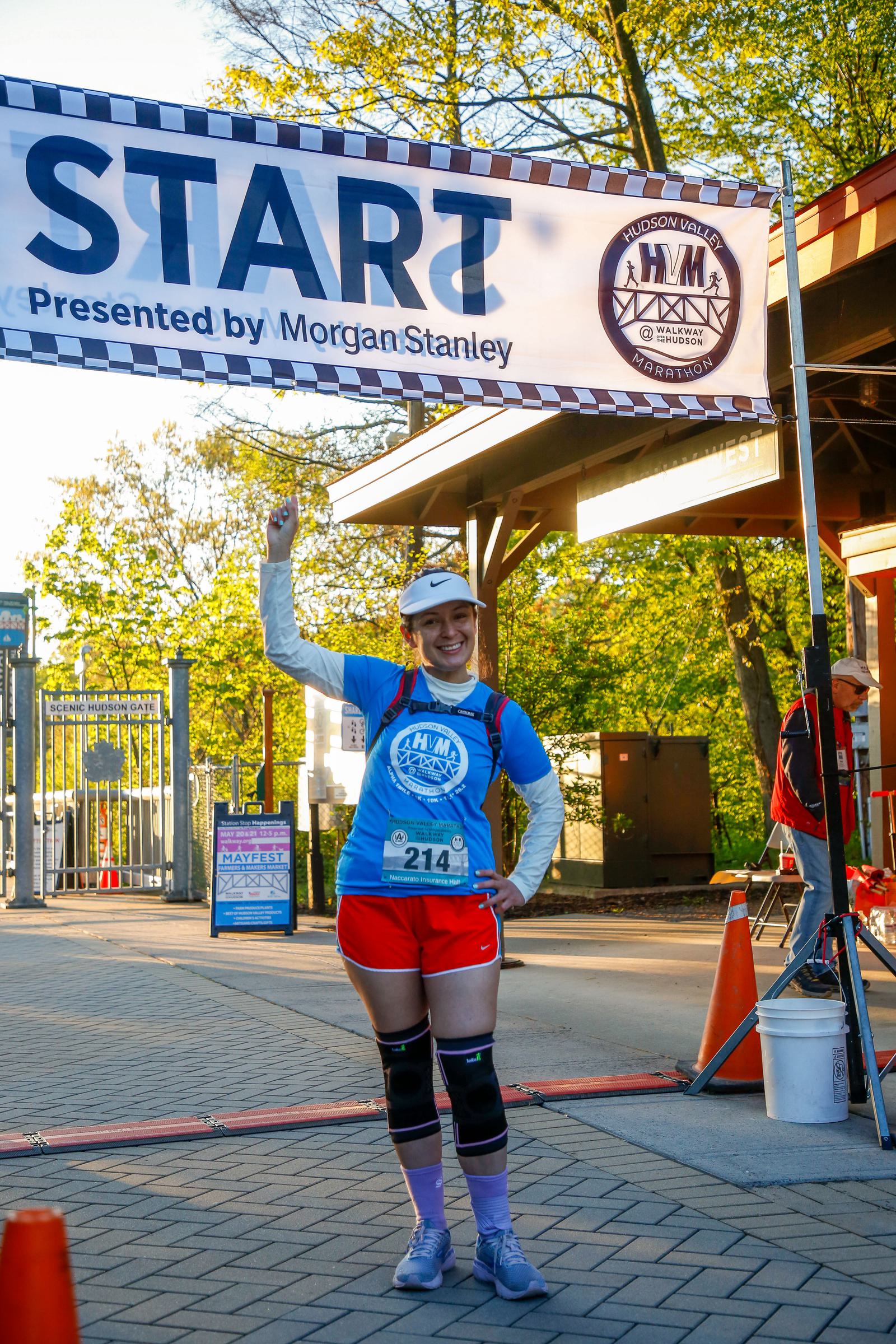 A person in athletic gear stands confidently under a "Start" banner at an outdoor event. They are wearing a blue shirt, red shorts, knee supports, and a white cap, with race number 214. The background features trees and a building.