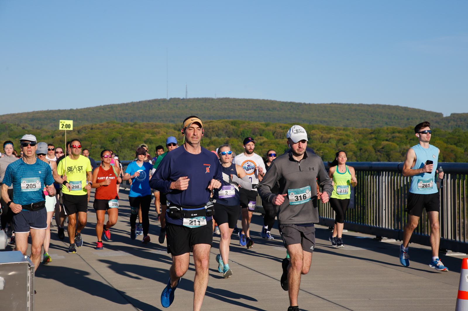 A group of runners competes in a race on a clear day, crossing a bridge with a green, hilly landscape in the background. Participants wear numbered bibs and a variety of athletic attire. A pacer holding a "2:00" sign is in the crowd.