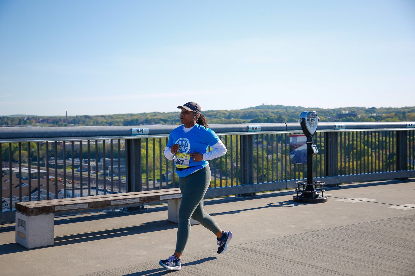 A person in athletic gear jogs on a bridge on a sunny day, with a scenic view of trees and distant hills in the background. A coin-operated binoculars stand is visible on the right side of the image.